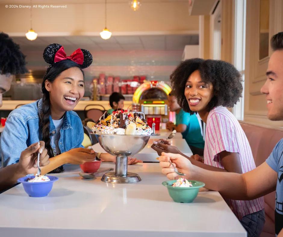 Beaches & Cream Soda Shop - Disney's Beach Club Resort 4 guests sat around a huge silver bowl full of many scoops of ice cream, whipped cream, sprinkles, m&m's brownies and cherries. One girl has sparkly minnie mouse ears on her head and both women are looking at one of the men. They are all holding spoons near their multicoloured mini bowls.