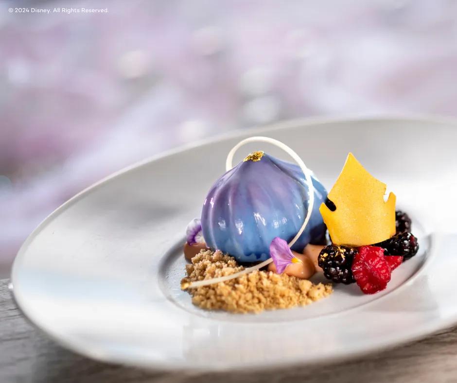 Topolino's Terrace - Disney's Riviera Resort A white plate of gorgeous food, what looks like a cheesecake from a fairy tale. There is a shiny blue and purple coloured tear shaped food on top of small crushed pieces of buscuit crumbs. There is a white chocolate arch over this and to the right there is a yellow crown shaped edible paper on top of raspberries and blackberries with small pieces of gold on them.