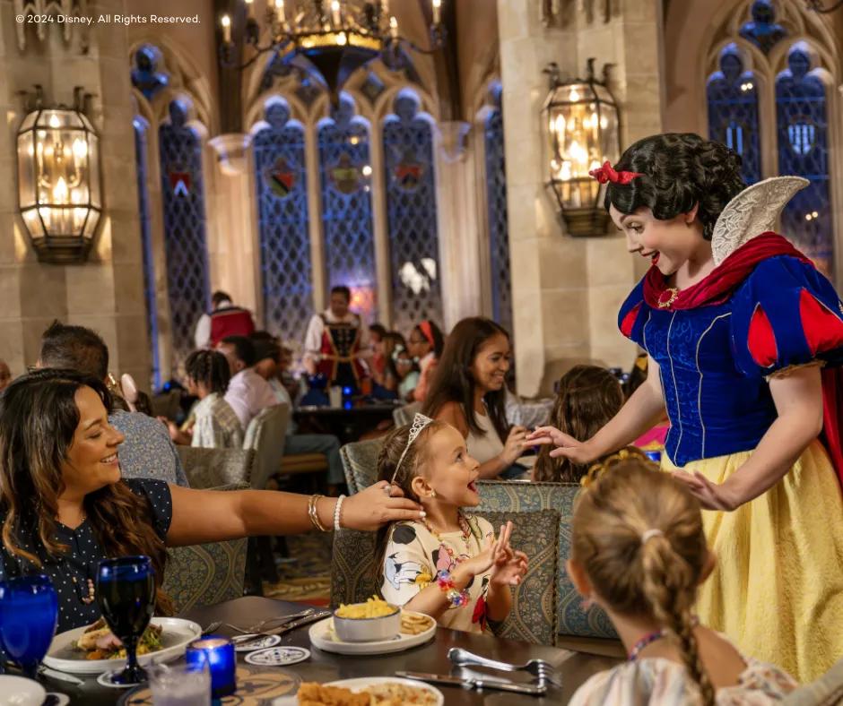 Cinderella's Royal Table - Magic Kingdom Park Princess dressed in blue and yellow dress with red details on it and a red cape as well as red bow in her black hair (Snow White) greeting guests at a table in a restaurant. A little girl looks up to the princess smiling and shocked with a tiara in her hair as her mother amiles watching, pulling back a strand of hair from the child's face. There is also an older sister looking at the princess with a plait in her hair.