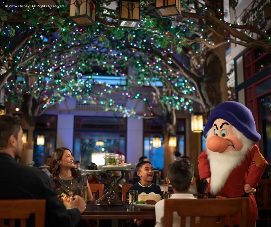 Artist Point - Disney's Wilderness Lodge A restaurant with dark lighting yet cosy, above there are decorative trees inside with multicoloured twinkling lights, below there is Grumpy the dwarf, a man with a large nose and furrowed eyebrows wearing a blue hat and a red outfit. Grumpy has his hands on his hips and is looking towards guests sat at a wooden table, a family of 4, a mum, dad and two boys who are smiling/laughing at him.