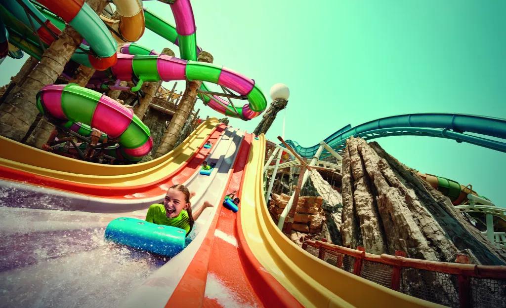 Yas Waterworld A huge pink and green stripey waterslide to the left, a large steep blue waterslide to the right and down the centre there is a racing waterslide where kids lay on stomachs on mats down small hills. There is a little girl winning, smiling with joy.