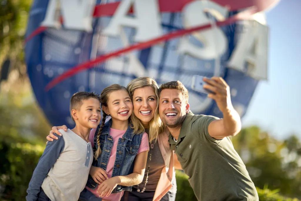 Kennedy Space Center Family of four, a man holding up a phone to take a selfie with wife and two kids, one boy and one girl. They are stood in front of a large sign, shaped like the earth, circular with red stripes over and the letters in white spelling NASA.
