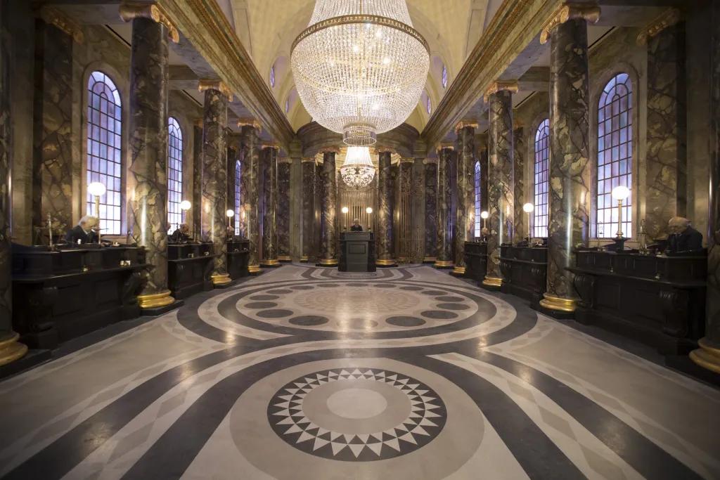 Harry Potter and the Escape from Gringotts Large clear building with black and white tiled patterned floor. Above hangs two grand chandeliers. Along the left and right hand side are tall dark brown desks with 3 goblin characters sat at them working. In the middle there is a larger desk with the main goblin working. This is a room themed as an elaborate bank.