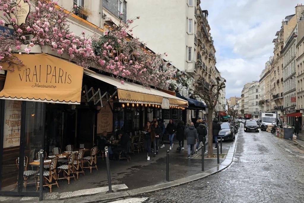 Paris restaurant A wet road in Paris lined with parked cars and the outside of s restaurant which is named "Vrai Paris". Above the beige cover there are pink flowers and below there are tables with chairs.