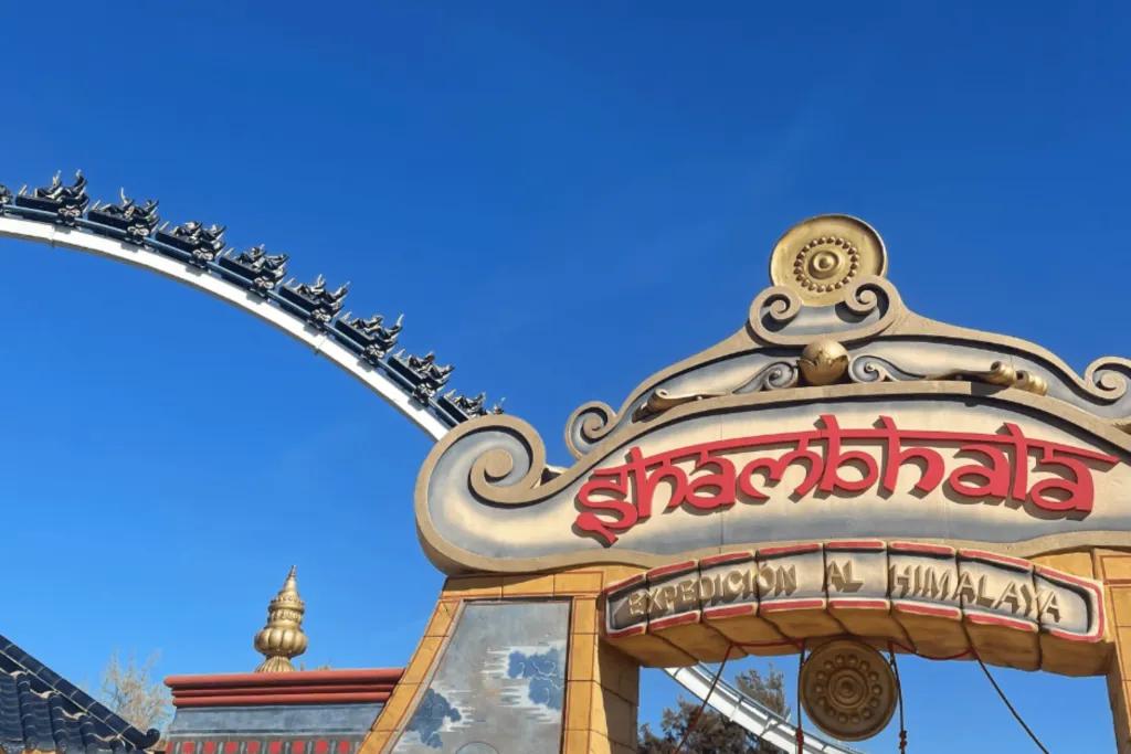 PortAventura Theme Park A large sign which is shaped as an archway which says shambhala - expedicion al Himalaya. The sign is coloured red and gold with patterns and artistic drawings of people on it. Behind there is a large white rollercoaster with a black rollercoaster carriage on it.