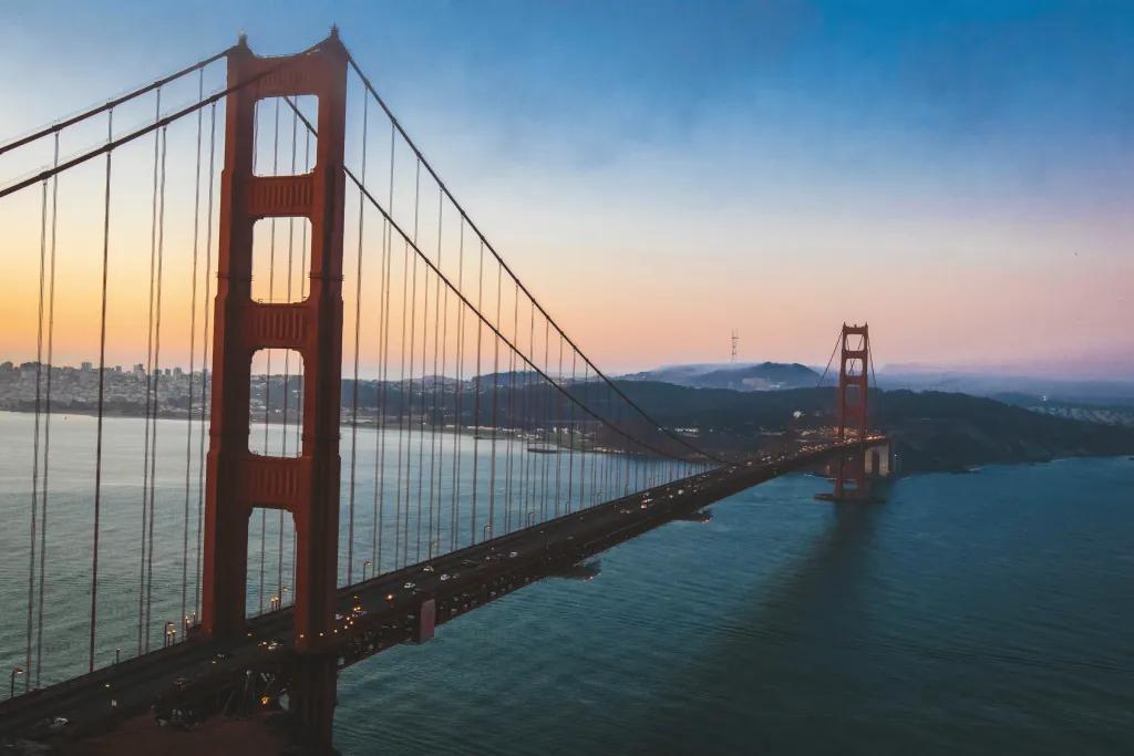 Golden Gate Bridge The sun is setting with blue and orange hues. Over a large river, there is a large red suspension bridge