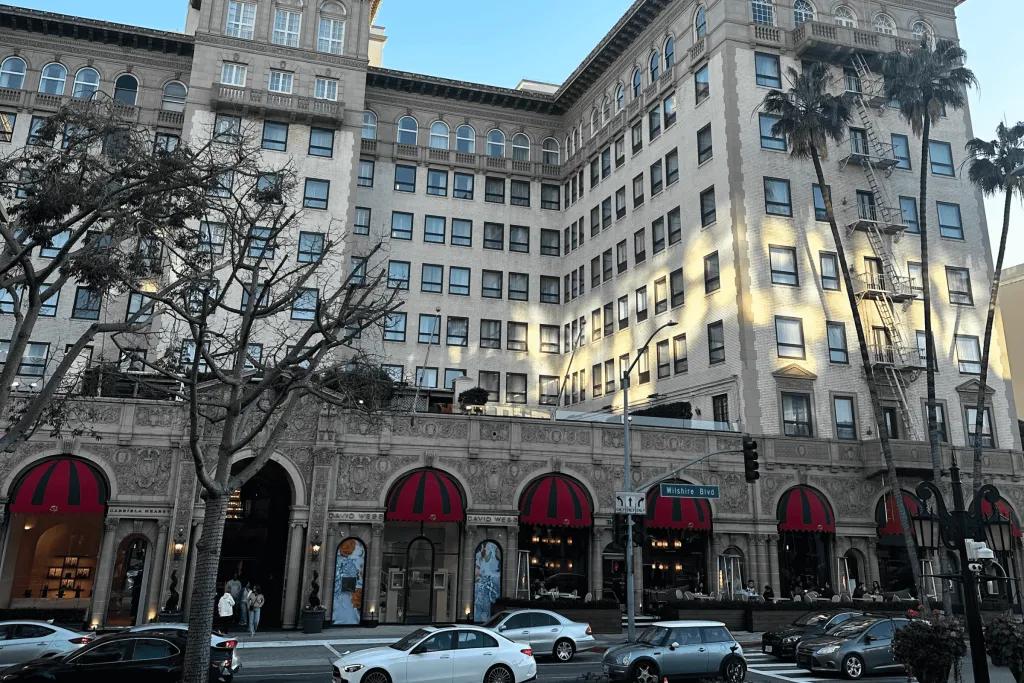 Beverly Wilshire, A Four Seasons Hotel A large clean building on the side of a busy road with palm trees in front of it. At the front of this building there are covers over windows coloured red and black.