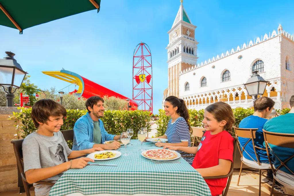 Ferrari Land A family of four, a dad, mum, son and daughter, all sat at table with green and white checkered table sheet eating pizza and pasta with a beige, white and green building behind them. To the left of the building there is a tall red rollercoaster with a Ferrari sign on the middle (yellow shield shape with black horse)