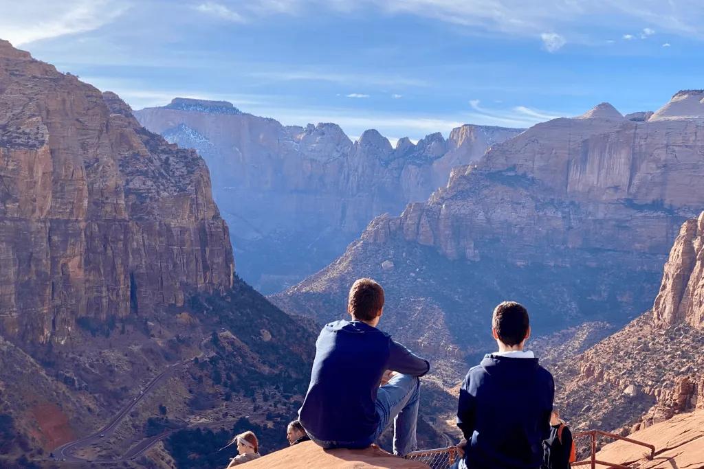 The Grand Canyon Two young men sat at the top of a large canyon. Slightly below there is a red railing with a couple of other tourists stood by. At the bottom of this large rock canyon there are trees and roads curving along the side.