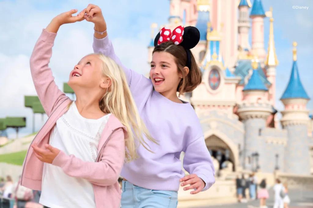Disneyland Paris A girl holding hands above head, twirling another little girl around in front of a large blue and pink castle. There are guests stood behind walking towards the castle.