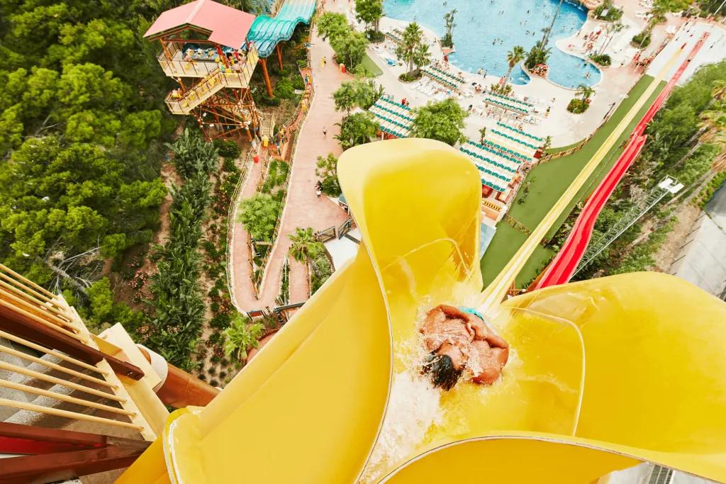 PortAventura Caribe Aquatic Park Birds eye view of man dropping down the top of a yellow vertical drop slide, beside there is a red which is smaller. There are pools below and walkways with people queueing for smaller waterslides.