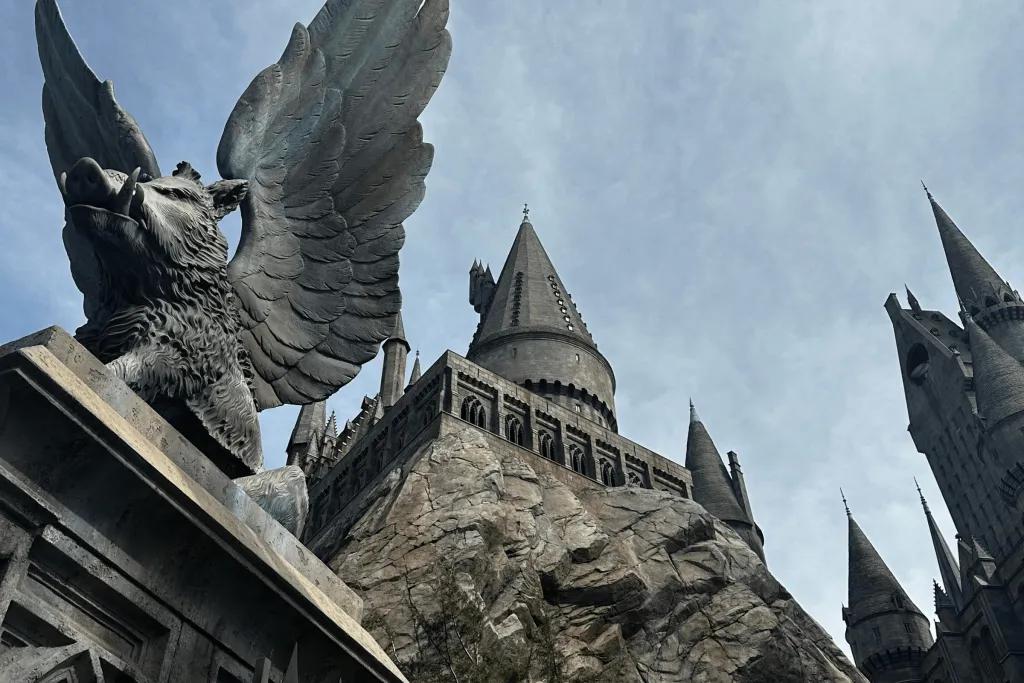 Hogwarts Castle Dark grey castle with pointed roofs sat on top of large rocks. To the left there is a statue of a pig with tusks and wings.