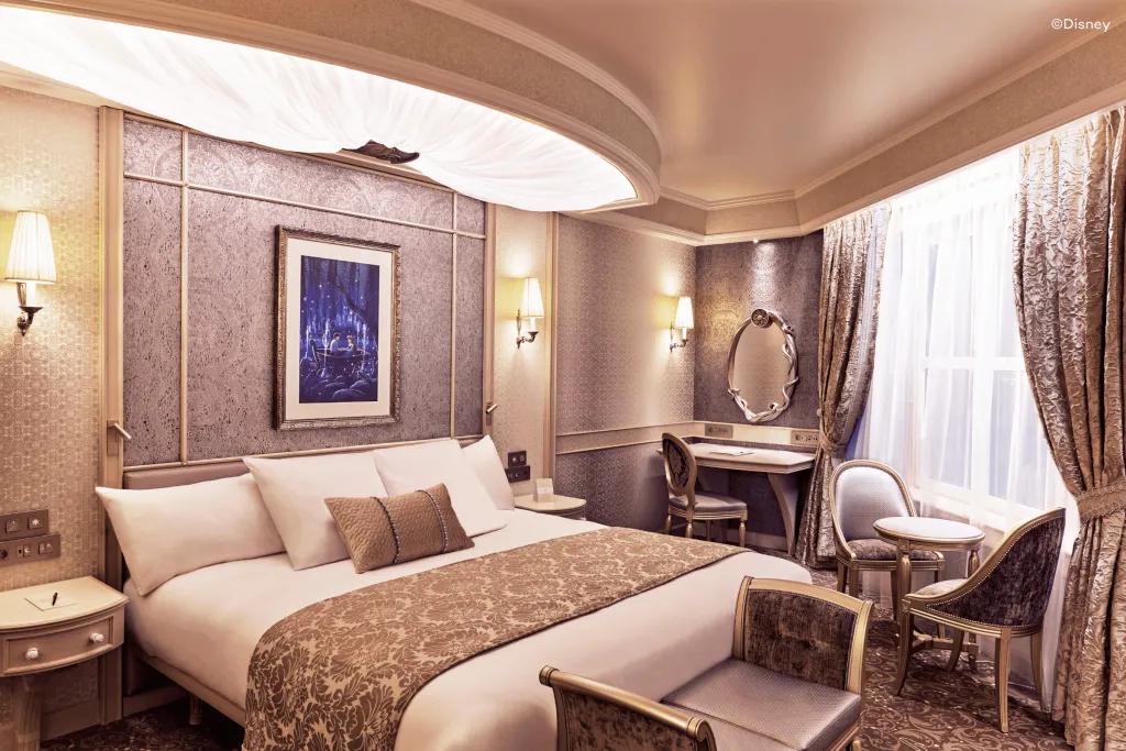Disneyland Hotel A bed with white and beige sheets and small cushions on. The room looks royal with a makeup table and a circular mirror above. There is a painting on the wall of Eric and Ariel in a boat holding hands.