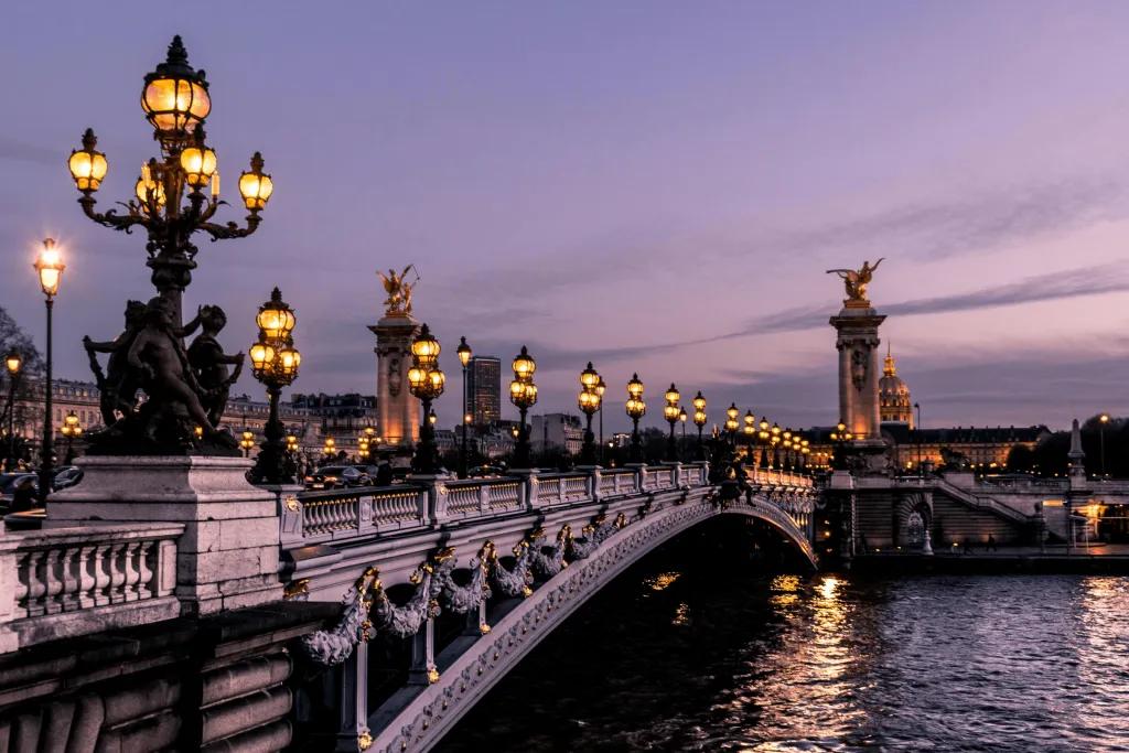 Paris River with bridge over lined with black lamp posts lit up. There are magnificent artistic golden and stone statues.