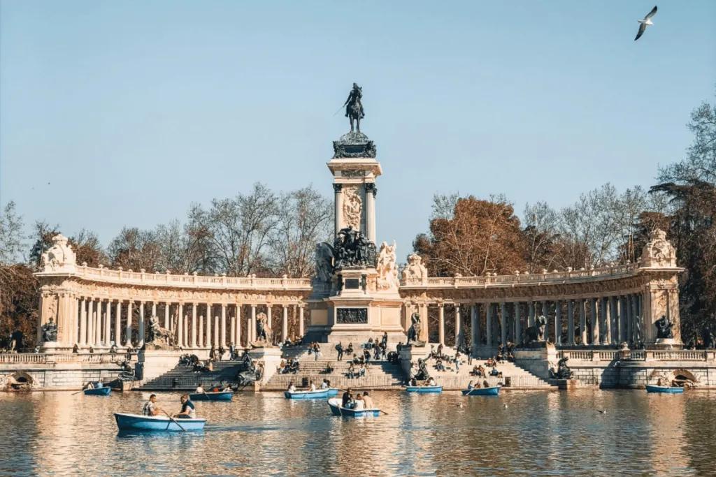 Madrid Large stone pillars lined up behind a stone statue, on the top there us a man sat on a horse. There is a lake in front of it with couples sat in blue rowboats.