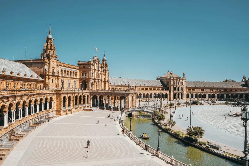 Seville Grand historic building in Seville which is beige coloured with white detailing. There are many archways along the side held up by white pillars. There are spires, and there is a wide white walkway, with a river running alongside it, with white bridges over the river.