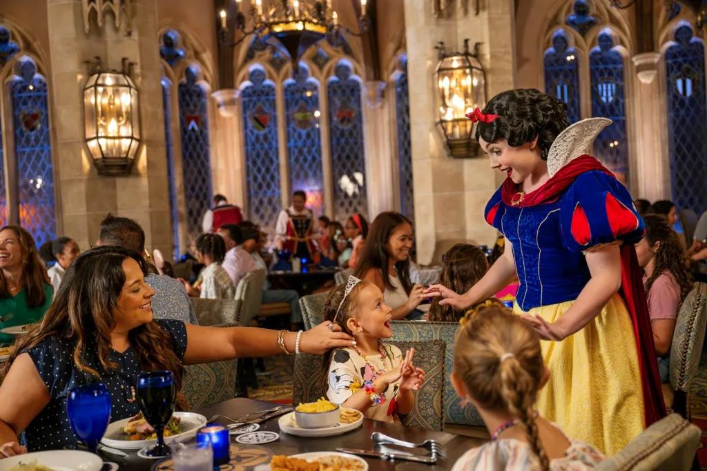 Cinderella’s Royal Table Mum and two girls sat a table eating. The mum is touching one of the girls hair as the little girl beams up at Princess Snow White who is at their table. Snow White is looking down smiling with a shocked expression. The small girl looks so surprised and happy. In the background there are tall windows on the brick walls and a large chandelier.
