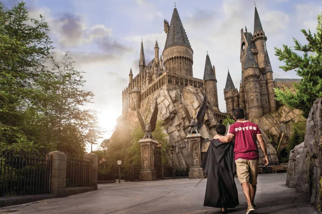 Hogwarts Castle A man wearing a red Griffindor shirt holding hands with a child in a black cloak and a wand. They are walking along an empty walkway in front of Hogwarts Castle.