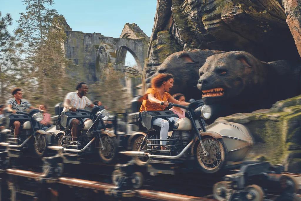 Hagrid's Magical Creatures Motorbike Adventure coaster Guests sat on motorbike shaped rollercoaster vehicles in front of stone walls and a three headed dog sat on rocks.