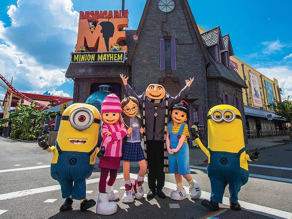 Despicable Me Minion Mayhem There is a black and grey house in the background with a large orange and yellow sign saying "Despicable Me Minion Mayhem" on. There are two minions in front with the Despicable Me characters (Gru, a man with a bald head, Agnes, a little girl with a black ponytail and dungarees on, Margo, a young brunette girl with glasses and Edith, a blonde child with a pink stripey hat on)
