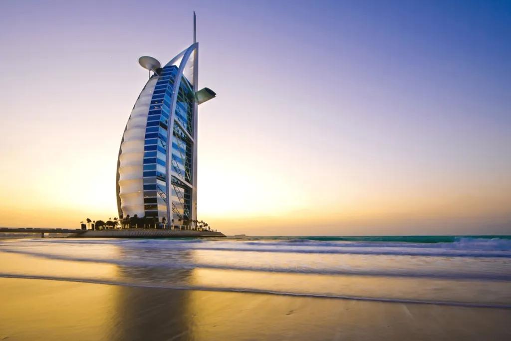 Burj Al Arab Sail shaped building coloured white with many windows and a helicopter pad on top. It is in the middle of the ocean on a man made island, with one road connecting it with mainland.