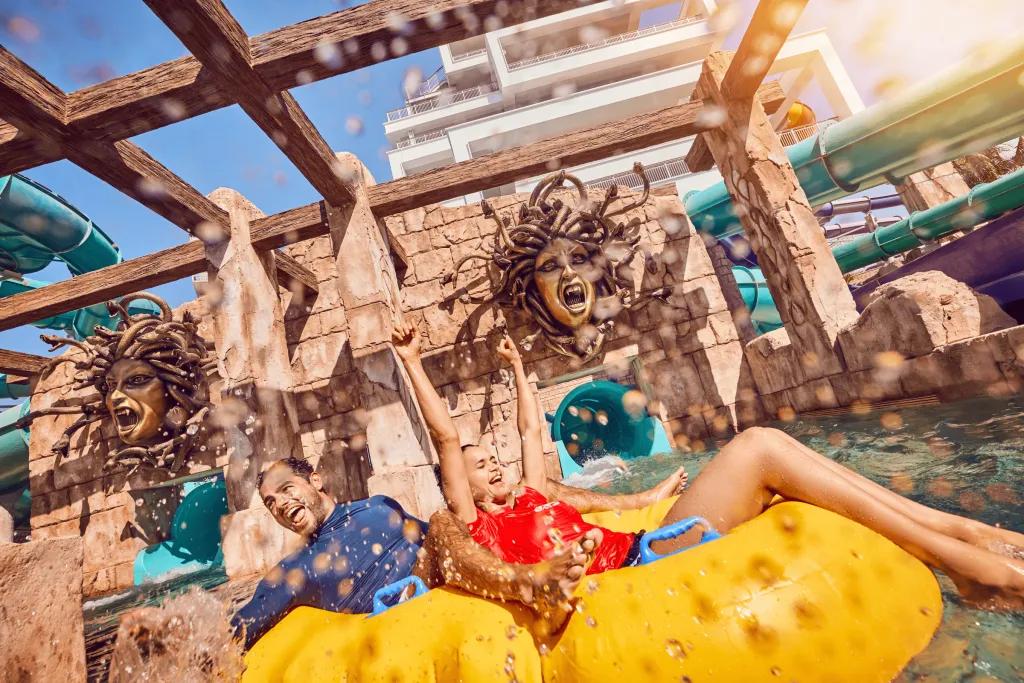 Atlantis Aquaventure Waterpark Man and woman in a double yellow rubber ring with water splashing around them. Behind them there are two waterslide exits coming out of a stone wall. Above there are golden face statues of Medusa.