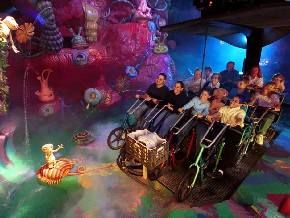 ET Adventure Ride A hanging ride vehicle with bicycle shaped seating. Guests are sat on these smiling whilst looking at the immaculately themed surroundings, yellow, pink and blue alien figures and flowers.