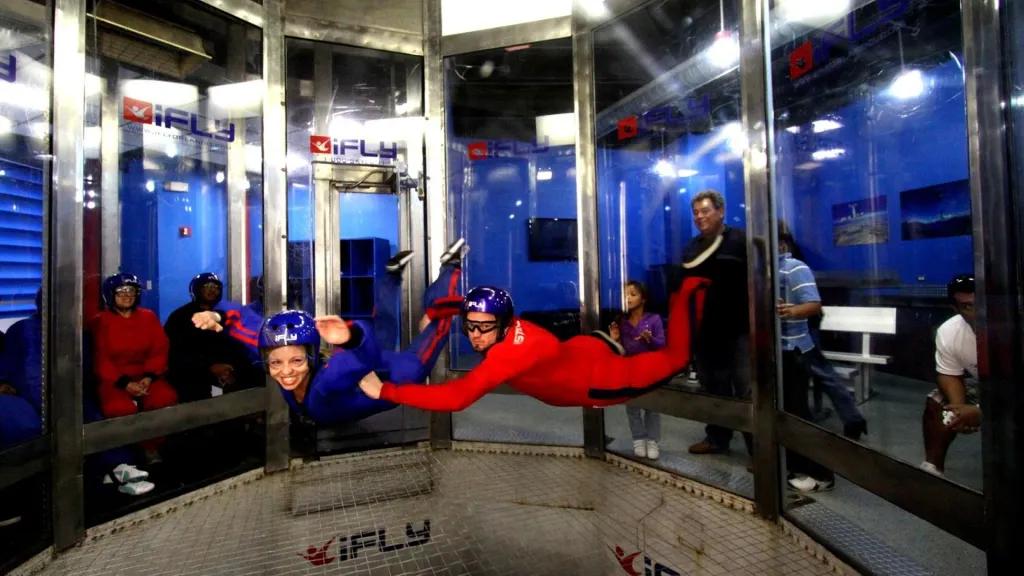 iFLY Orlando A large fan with windows all around inside a blue building and people peering in watching as a man in a full red jumpsuit, a helmet and goggles holds onto a girl wearing a blue jumpsuit, a helmet and goggles as the both float in starfish positions.