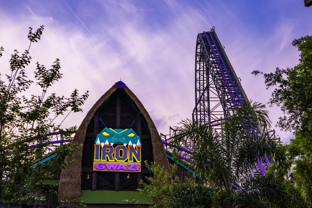 Iron Gwazi in Busch Gardens Tampa Bay Iron Gwazi sign with green snake cartoon on top. In the background there is a tall purple roller coaster hidden behind trees and the sky is also a pink/purple shade.
