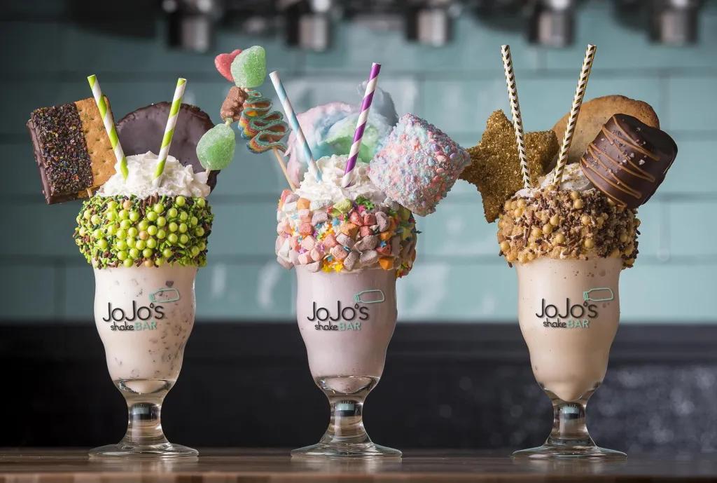 JoJo's Shake Bar - Pointe Orlando Three huge milkshakes in glasses lined up, one with green balls lining the top of the glass, a pink drink and two large biscuits sticking out with a cocktail stick of sweets and two green straws. The second one has pink and orange sweets lining the glass, a pink liquid with whipped cream, a giant marshmallow on a stick, pink and blue cotton candy and a stick with sweets on. Finally the last one has brown chocolate balls on the top of the glass, a chocolate flavoured milkshake and biscuits.