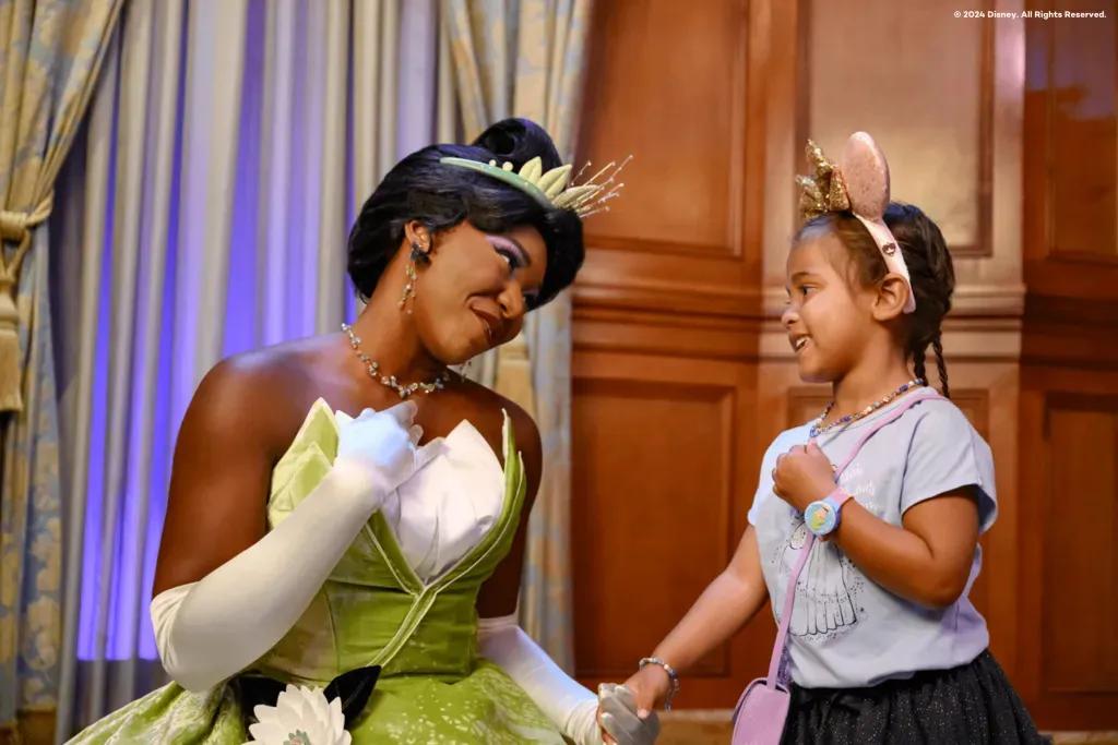 Prinzessin Tiana in der Princess Fairytale Hall Ein junges Mädchen mit rosa Minnie Mouse Ohren hält Händchen mit Prinzessin Tiana