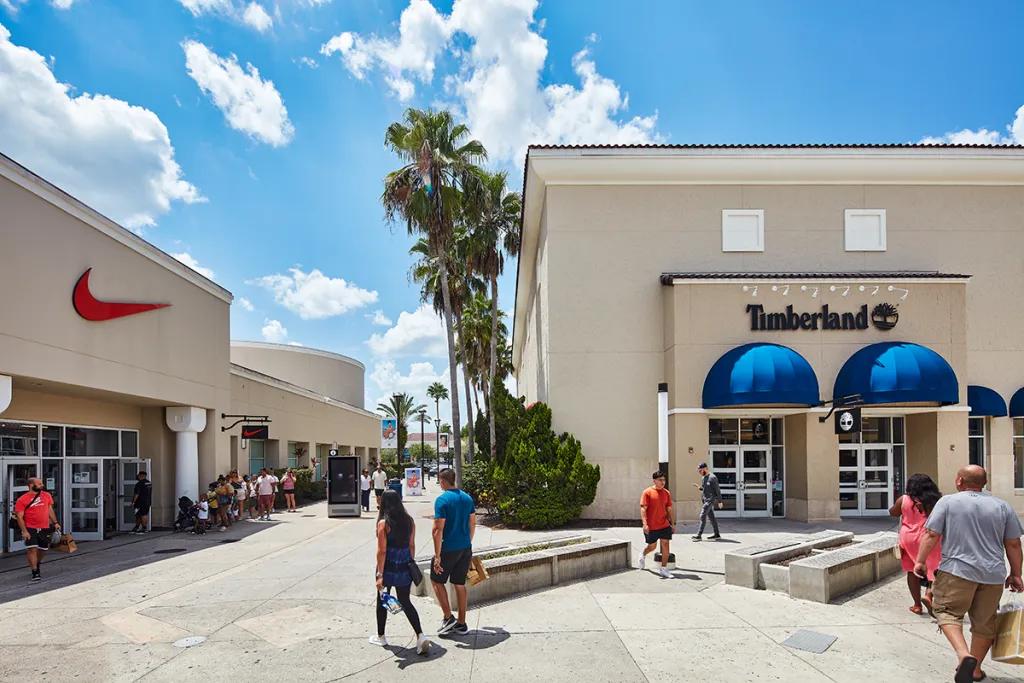 Vineland Premium Outlets People walking through rows of shops on either side lined with palm trees. People are holding shopping bags and there are shops such as Timberland and Nike.