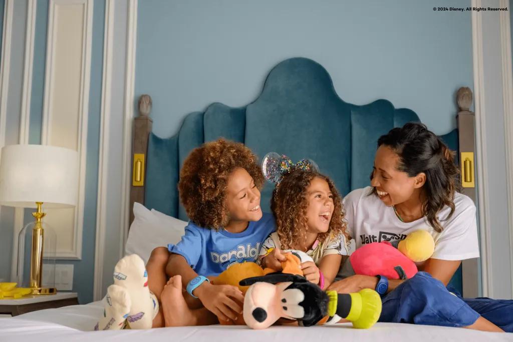 Walt Disney World Resort in Florida Resort A woman with her son and daughter sat on a bed with white sheets and a blue headboard with mickey ears on and each of them are clutching onto a plush toy of Mickey Mouse and Goofy.