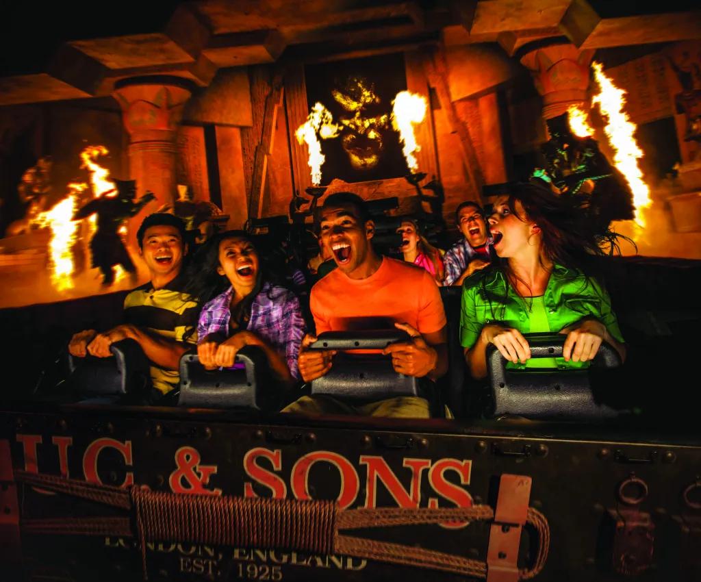 Revenge of the Mummy in Universal Studios Florida Four guests smiling and gasping as they clutch to their lap bars on a indoor rollercoaster. There is fire pyrotechnics surrounding them and silhouettes of mummy's jumping out on them.