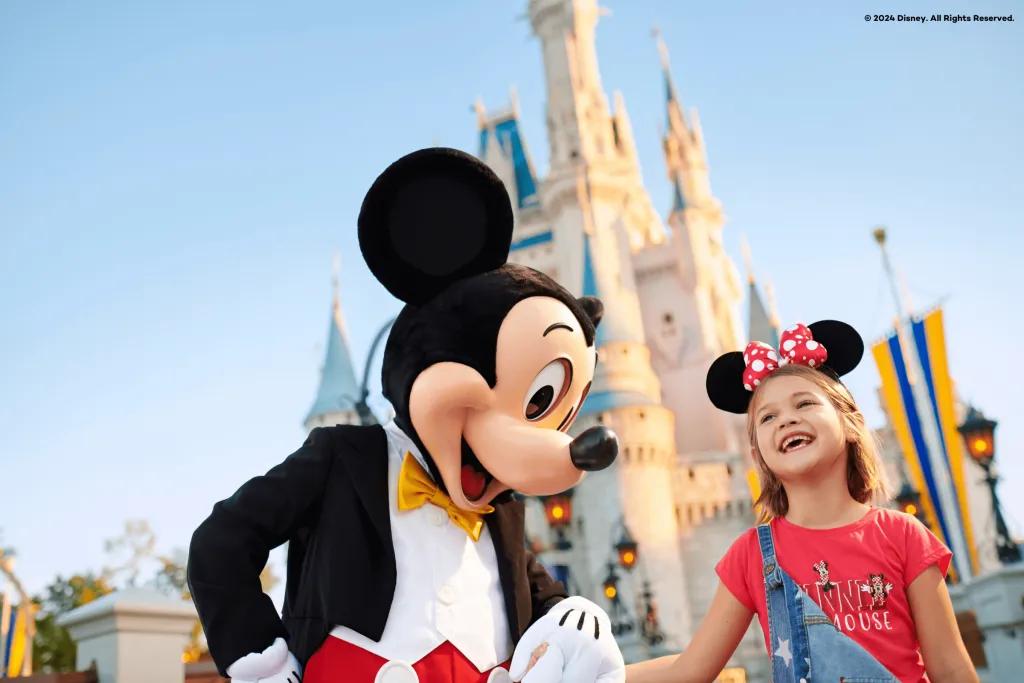 Magic Kingdom Park Mickey Mouse, a cartoon mouse wearing a yellow bow tie and a suit coloured red and black, holding hands with a young girl wearing Minnie Mouse ears stood in front of a white and blue castle at sunrise.
