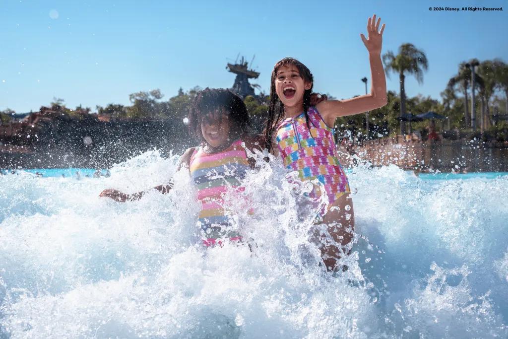 Disney's Typhoon Lagoon Two children smiling and laughing as they run out of a large pool with their hands in the air. There is water splashing around them and there is a statue of a boat stranded on top of a large rock in the background.