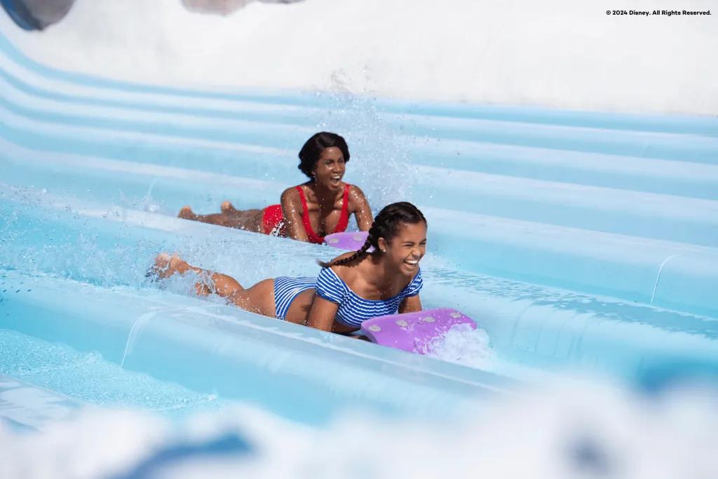 Disney's Blizzard Beach There is a mum and daughter sliding on mats down a light blue waterslide side by side. There are seven in a row for people to race down. Both are smiling.