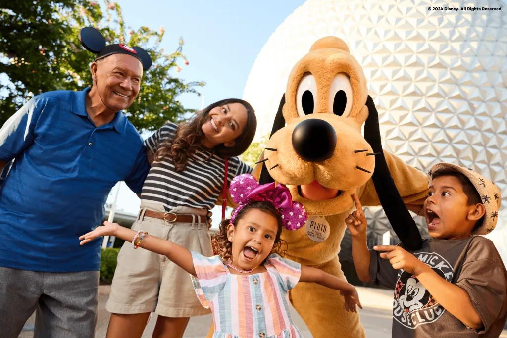 EPCOT There is a golf ball white shaped building in the background. There is a yellow cartoon dog called pluto stood with a family of four. The young boy is gasping pointing at his sister in shock, the daughter is stood in front with pink sparkly ears with her arms extended out, and the parents are stood arm in arm smiling.
