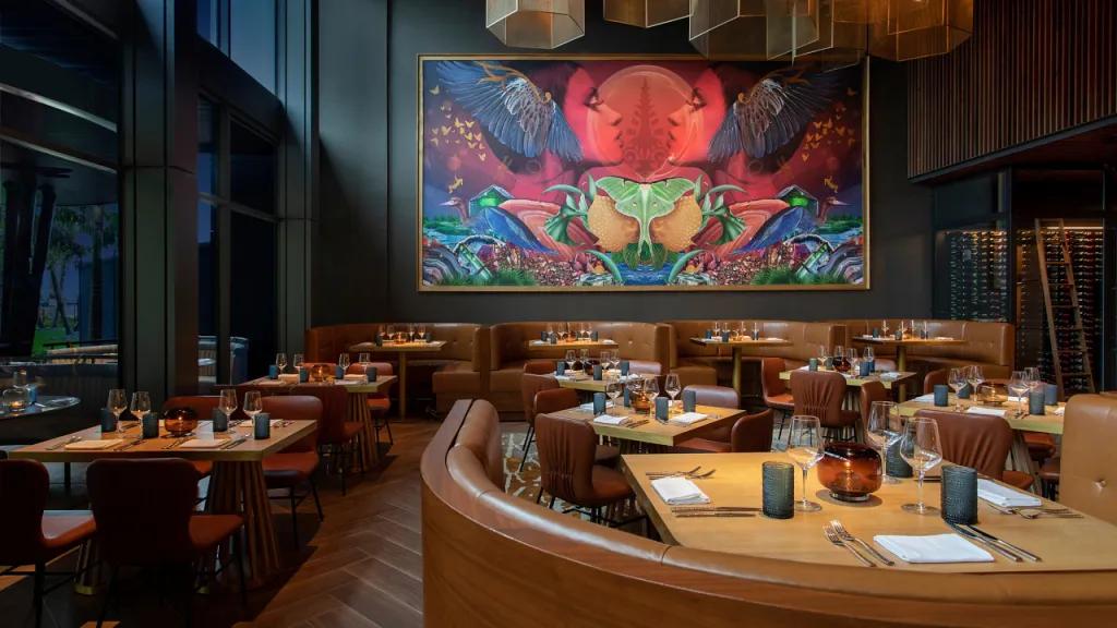 BACAN Dimly lit restaurant with round booths and large tables. There is a bright piece of artwork on the wall which is bright red, blue and purple which stands out amongst the dark wooden