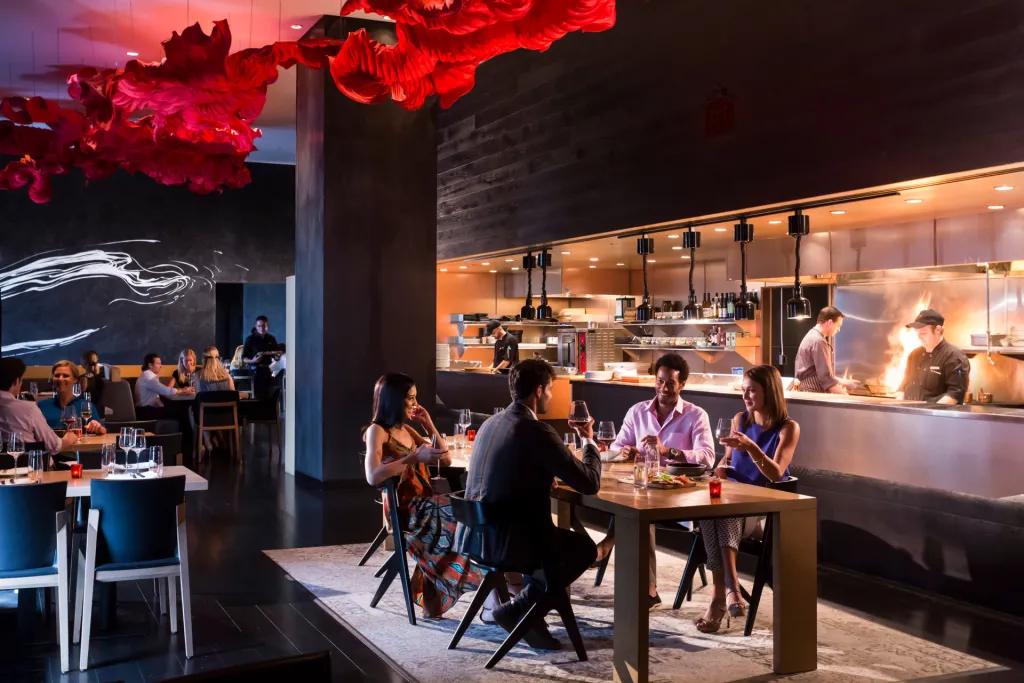 Capa Steakhouse & Bar Modern restaurant with four people sat around a table talking. Above there is an artistic reg light shaped like a cloud. Behind there is a kitchen with chefs and flames coming from frying pans.