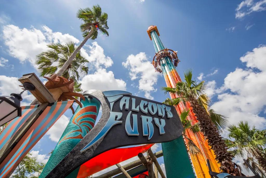 Falcon's Fury at Busch Gardens Tampa Bay A sign which says Falcon's Fury and there is a ride in the background which is a large drop tower with the ride in motion and the circular seating dropping through the sky.
