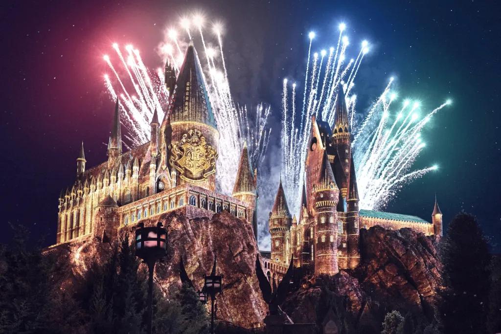 Hogwarts Always Large brown castle sat on top of rocks with many towers. It is night time and there are fireworks shooting out behind the castle of different colours, red, yellow, blue and green to match the house colours. There are also projections on the castle with it lit up yellow and a large shield with four different animals on it.