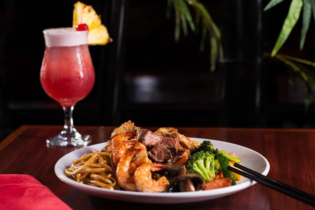 Kobe Japanese Steakhouse White plate with steak, prawns, noodles and broccoli on it mixed together. There is also a cocktail full of red liquid and a slice of pineapple on the glass.