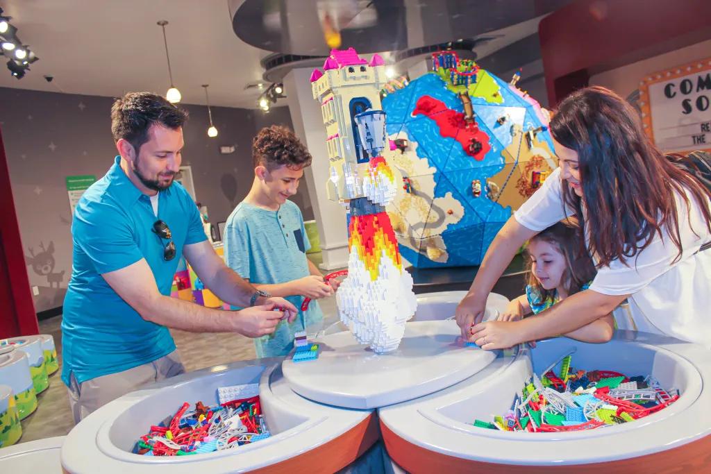 LEGOLAND Florida Resort Family of four crowded around a lego table building their own creations. The mum and daughter are stood together and the dad and son are stood on the opposite side. In the background there is a lego earth.