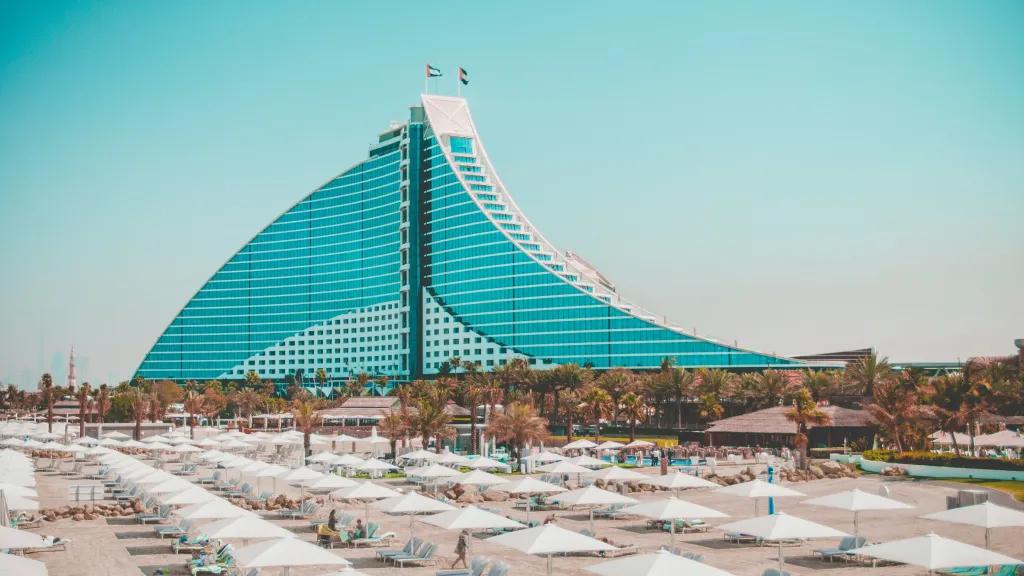 Jumeirah Beach Hotel Building shaped like a sail made purely of windows. On top there are two UAE flags, below there is a beach with pristine white sand and white umberellas.