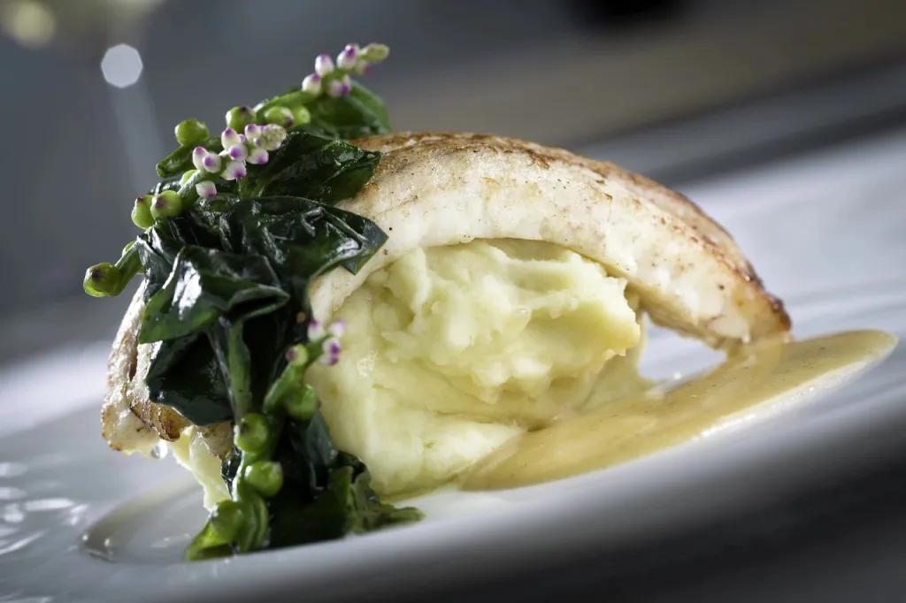 Norman's Slice of fish placed on mash potato scoop. There's green vegetables on top and a beige coloured gravy.