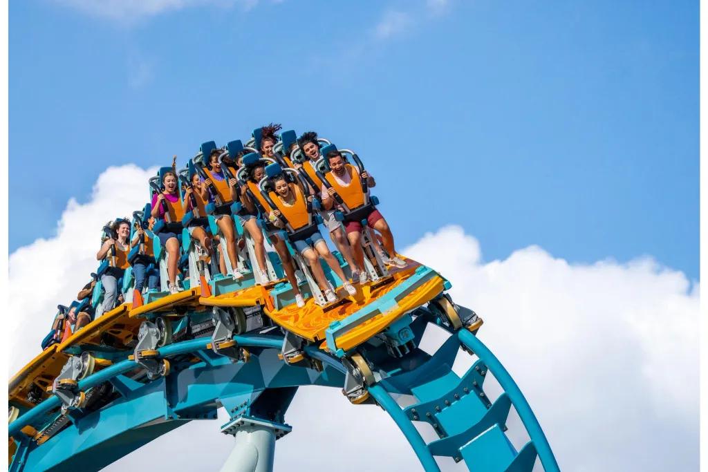 Pipeline in SeaWorld Orlando People on a rollercoaster car standing up with harnesses around their shoulders. They are stood on a vehicle which is themed as a surfboard coloured orange and turquoise with the track a light blue.