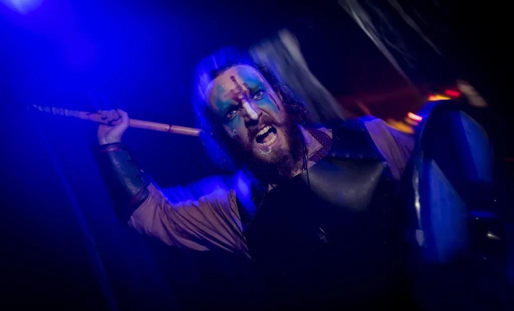 Scare Zones  A dark and blurry image of a man screaming whilst holding an axe which he looks like he's about to hit the camera with. He has face paint on and an apron.