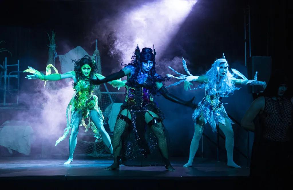 Sirens Song Three women dressed as sirens in blue lighting, one green, one black and blue and the other icy blue. They are wearing textured clothes and have matching hair. Each of them are in the middle of a dance with their arms out reached.