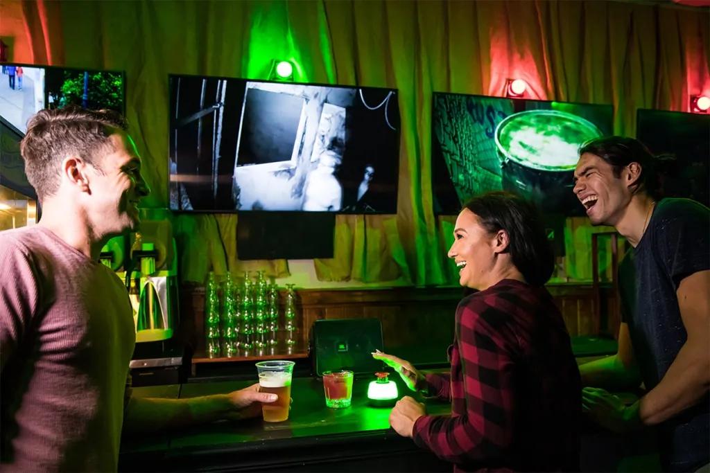 Themed bar Room with curtains and green and red lighting above TV's which look like they are playing CCTV footage. There are three adults sat with drinks all laughing with one woman holding her hand above a red button with white lighting around it.